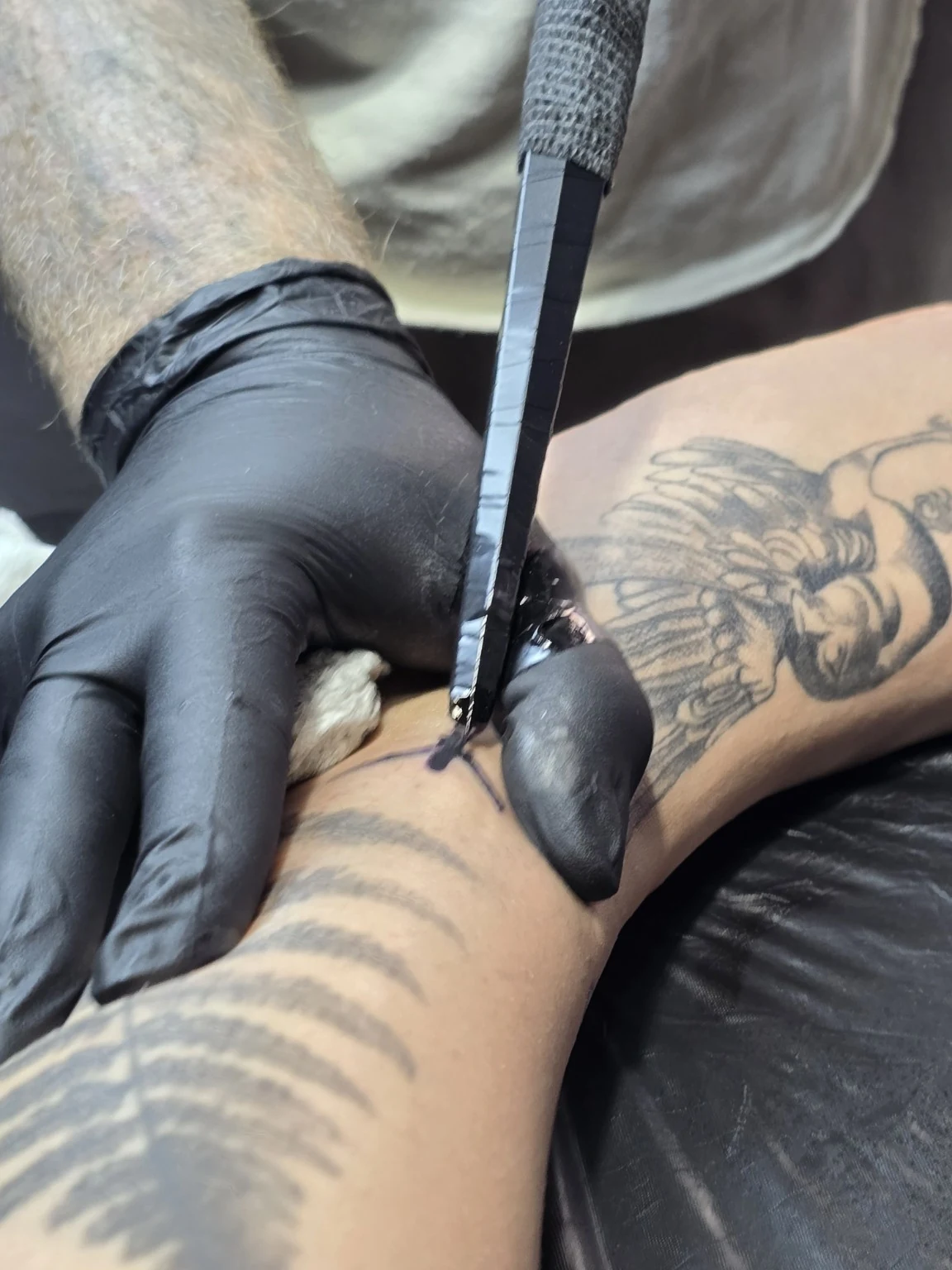 Gloved hands holding a tattoo tool against skin beside existing tattoos.