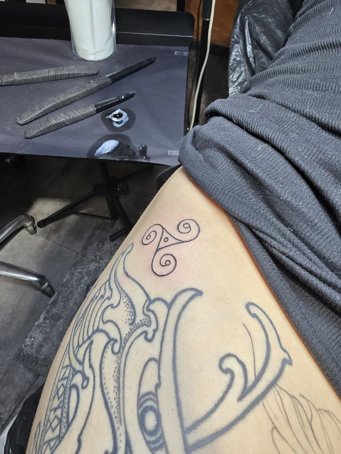 A small spiral motif on skin beside a larger existing outlined tattoo.